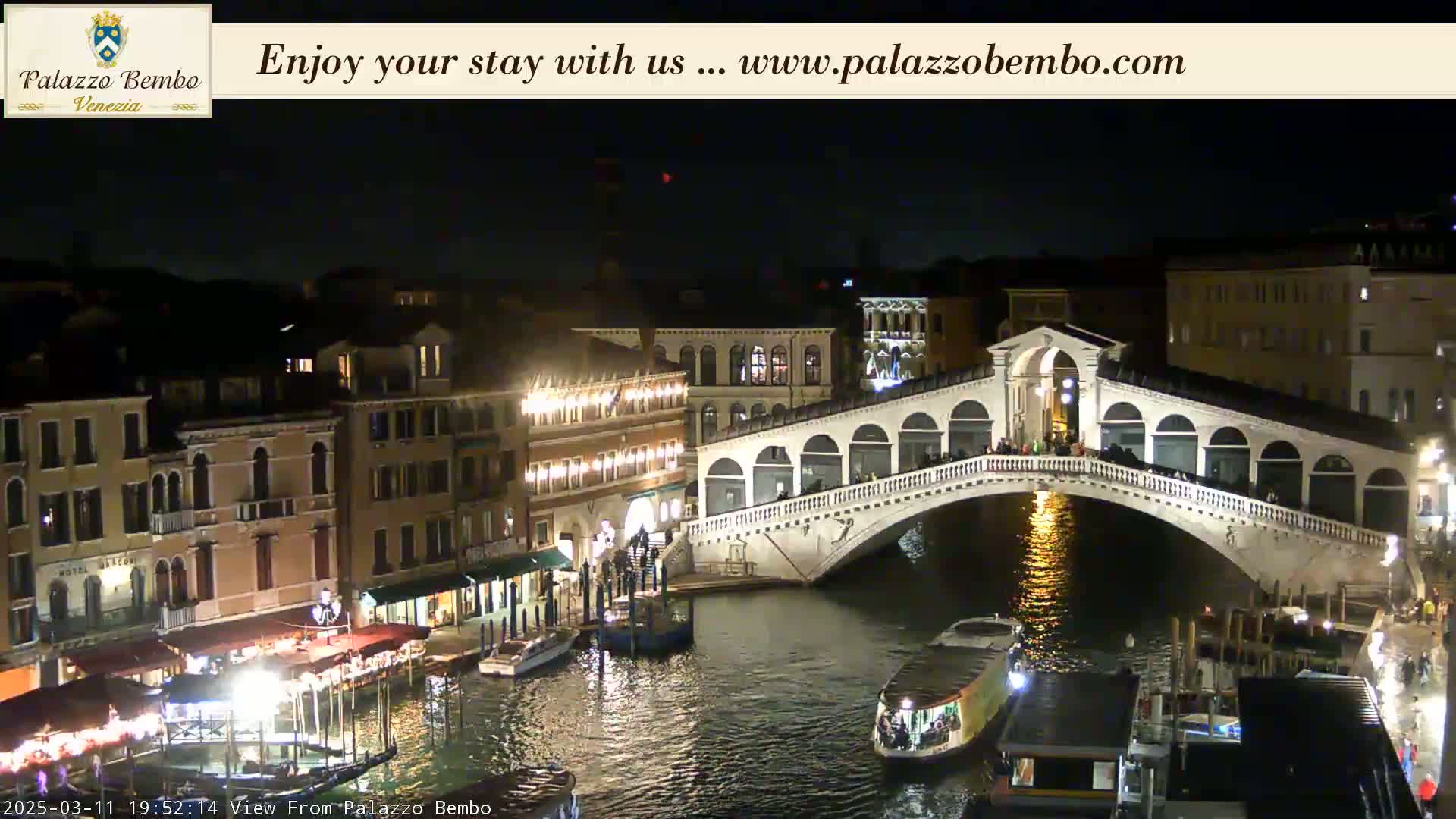 A nighttime view of a canal in Venice, showing a bridge with people on it, boats moored along the canal, and illuminated buildings lining the waterway.