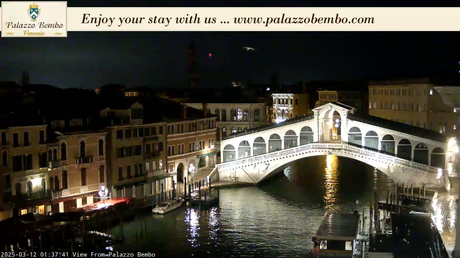A nighttime view of a canal in Venice, featuring a lit bridge and buildings along the water's edge under a dark sky.