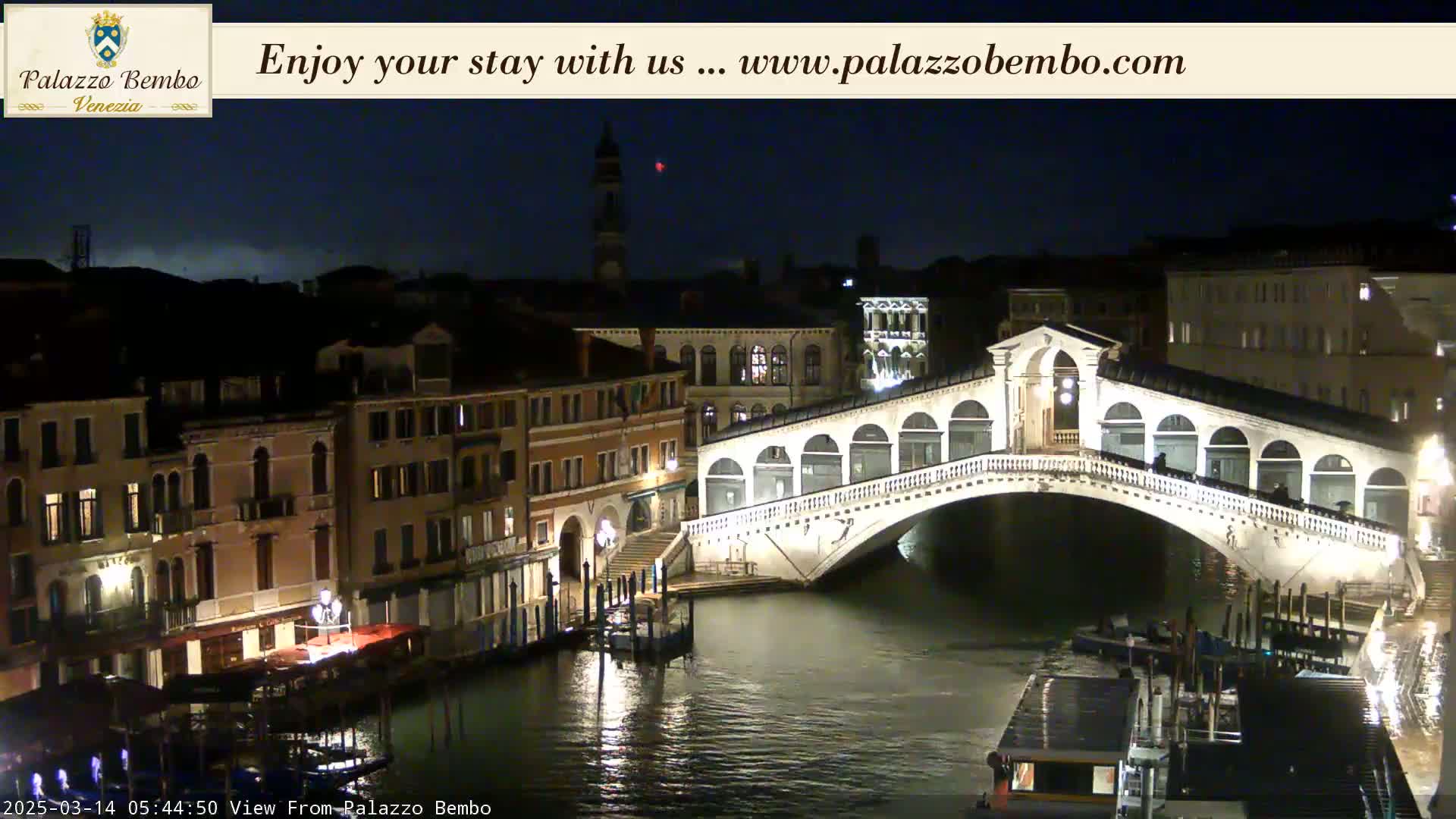 A nighttime view of a canal in Venice, showing a bridge illuminated against a dark sky.