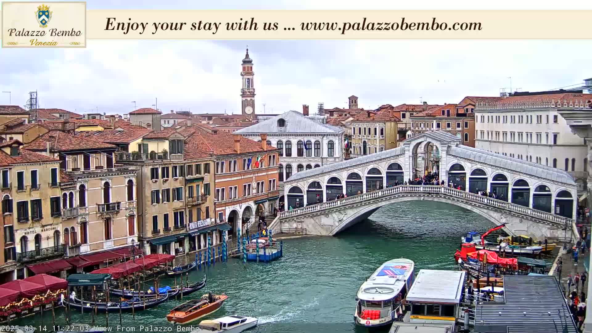 A view of a canal in Venice, under an overcast sky, shows a bridge with people on it and boats on the water, with buildings lining the canal.