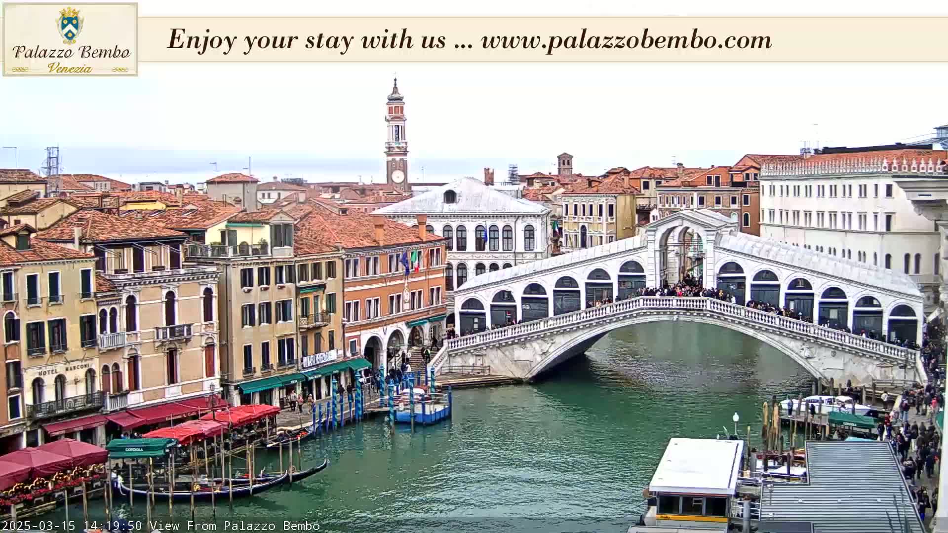 A canal in Venice, under an overcast sky, shows the Rialto Bridge crowded with people, with gondolas and other boats moored along the canal's edge.