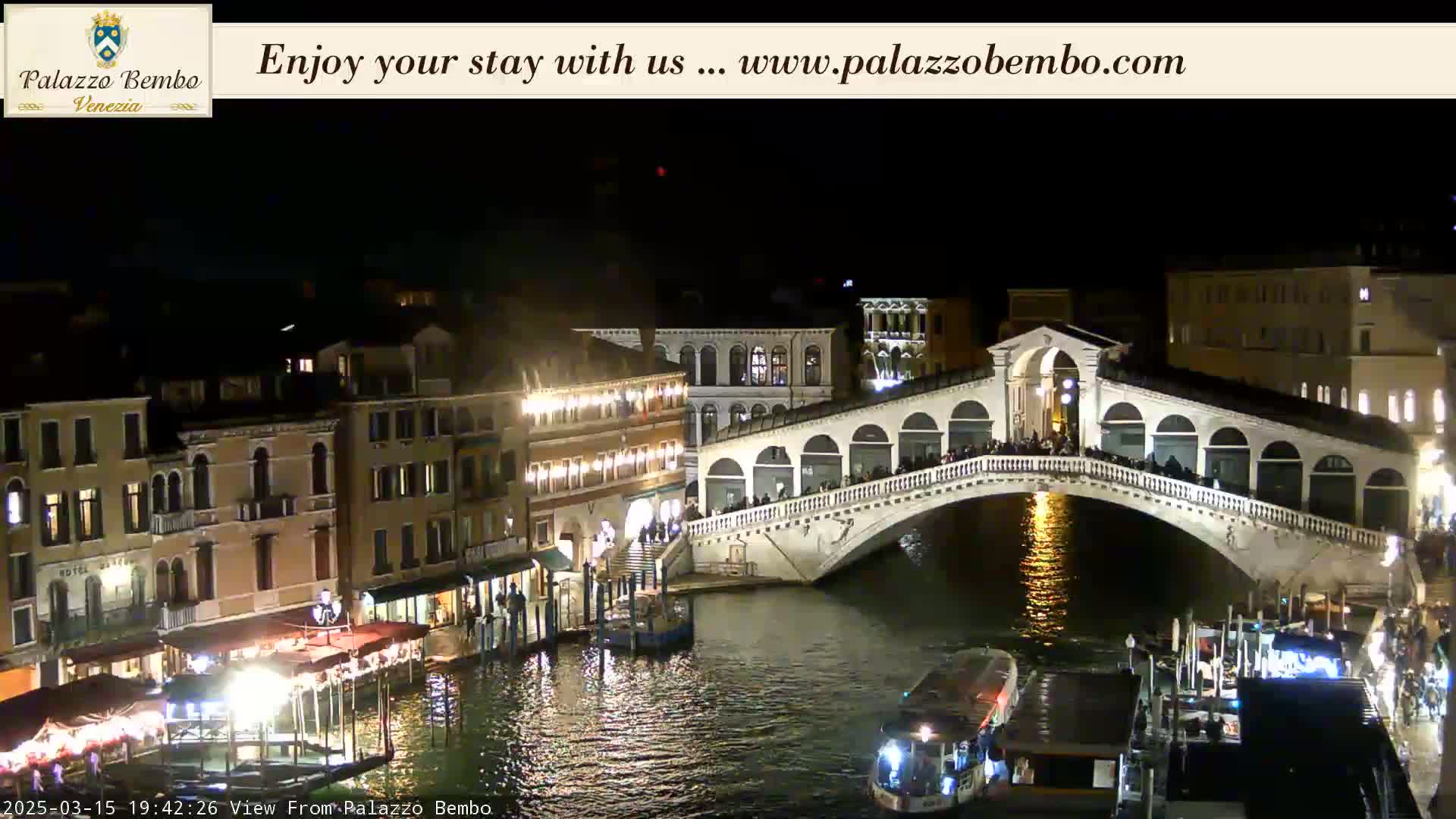A nighttime view of a bridge over a canal in Venice, with illuminated buildings and boats along the waterway.