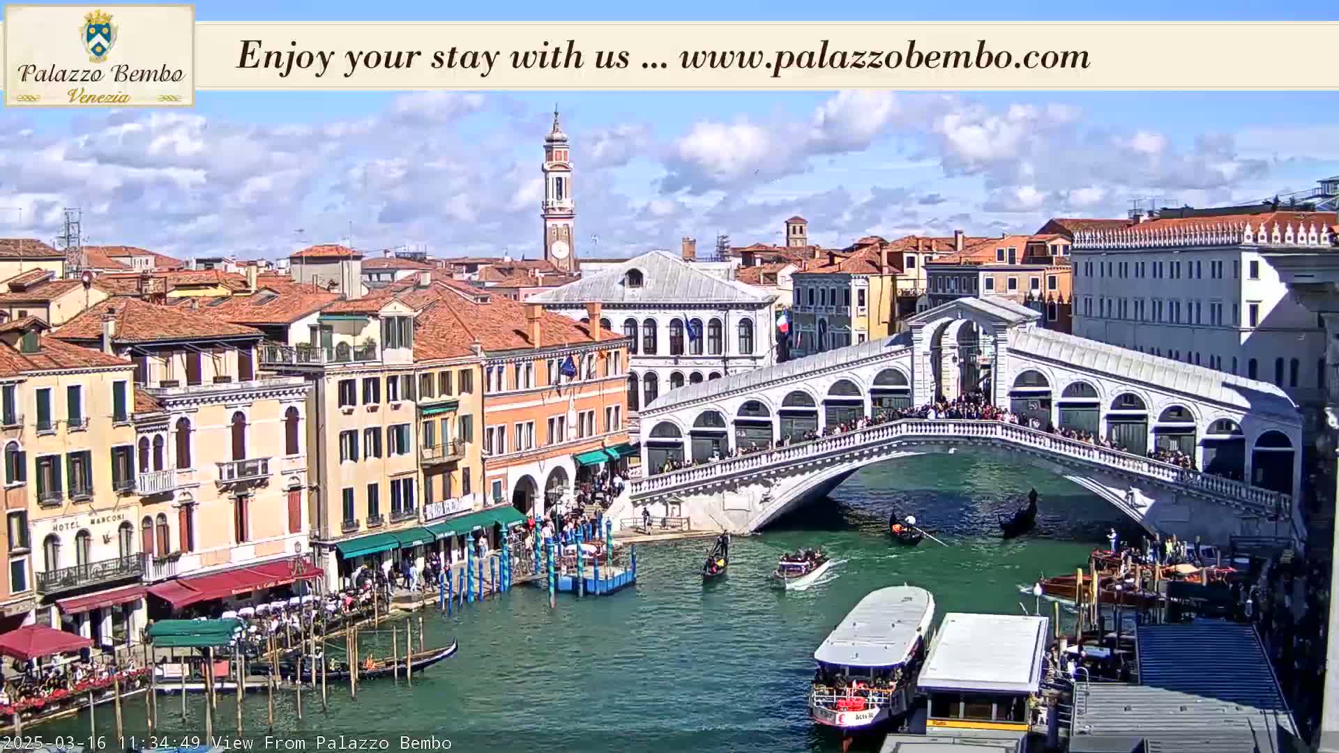A sunny day in Venice shows a canal with gondolas and water taxis, a bridge crowded with people, and numerous buildings along the waterway.