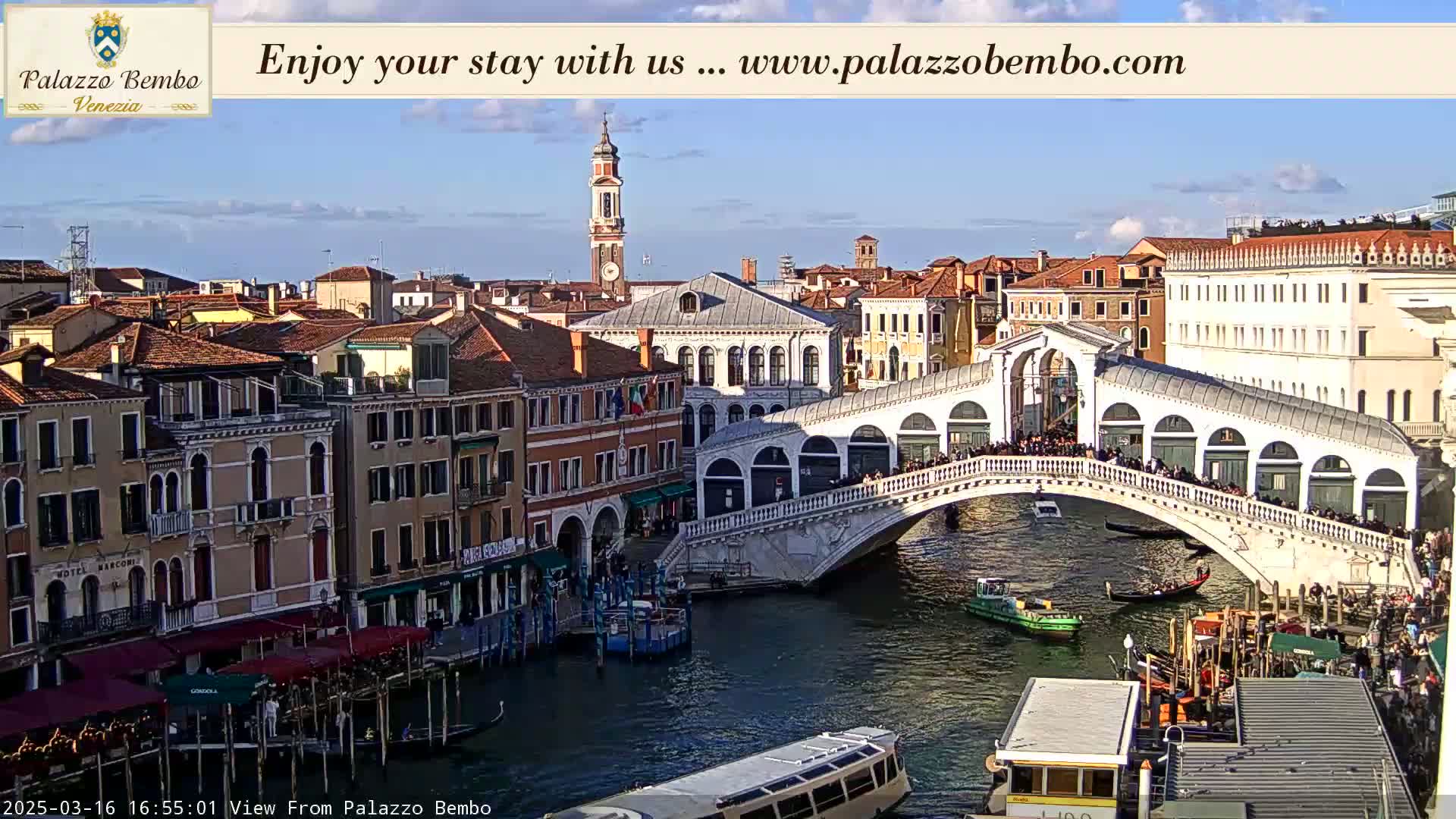 A sunny view of a canal in Venice, with a bridge crowded with people, boats on the water, and buildings lining the waterway.