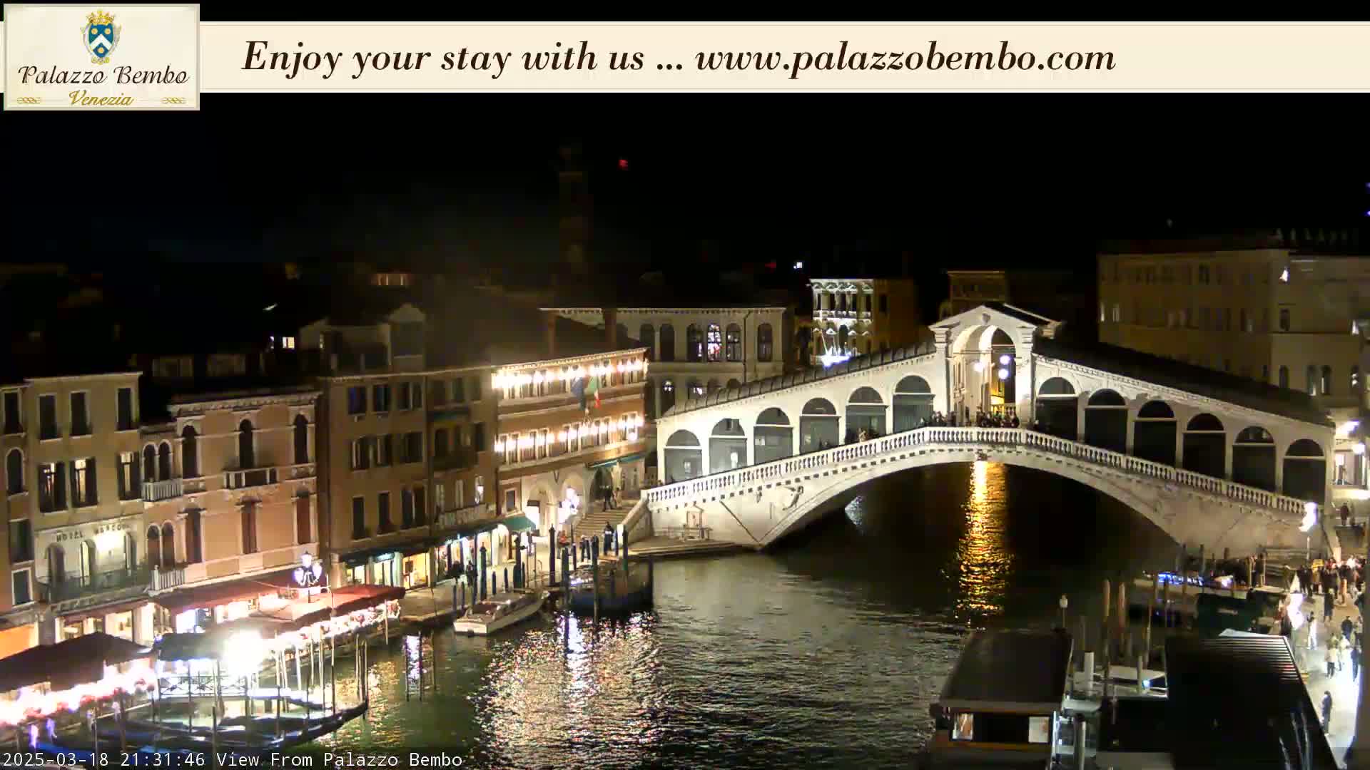 A nighttime view of a canal in Venice, showing a lit bridge and buildings along the waterway under a clear sky.