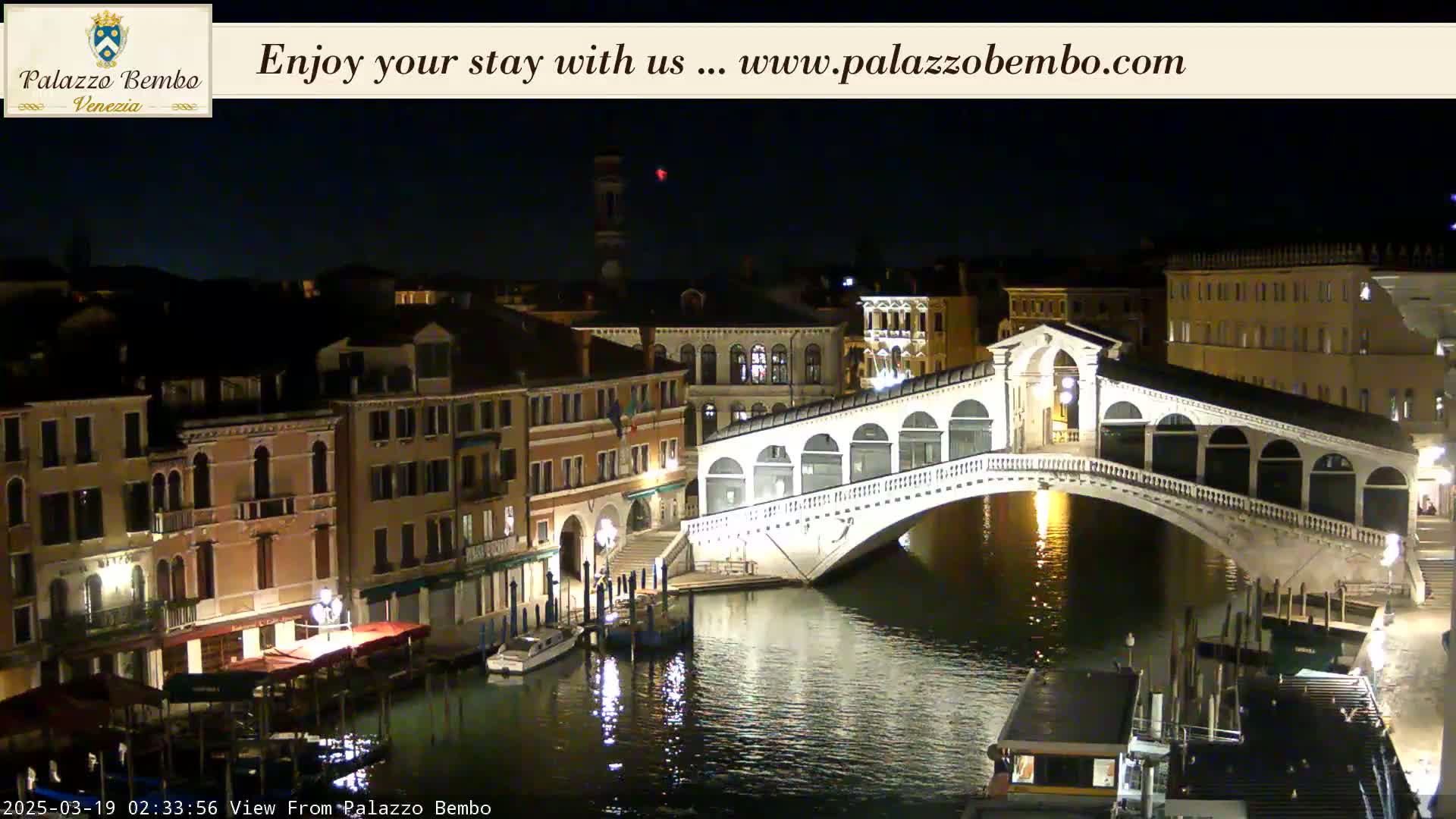 A nighttime view of a canal in Venice, showing a bridge illuminated against a dark sky.