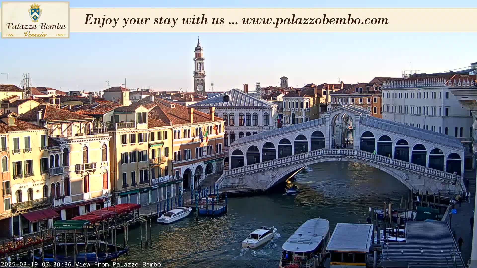 A sunny view of a canal in Venice, Italy, shows a stone bridge arching over the water, with boats and buildings lining the waterway.
