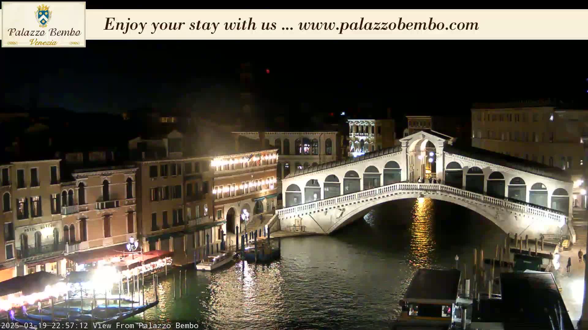 A nighttime view of a canal in Venice, showing a bridge lit up with lights, buildings along the water's edge, and boats moored nearby.