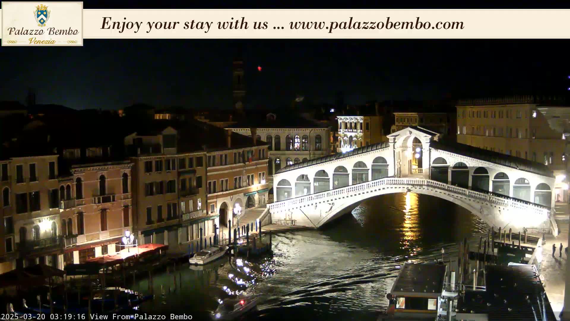 A nighttime view of a lit-up bridge over a canal in Venice, with buildings lining the waterway.