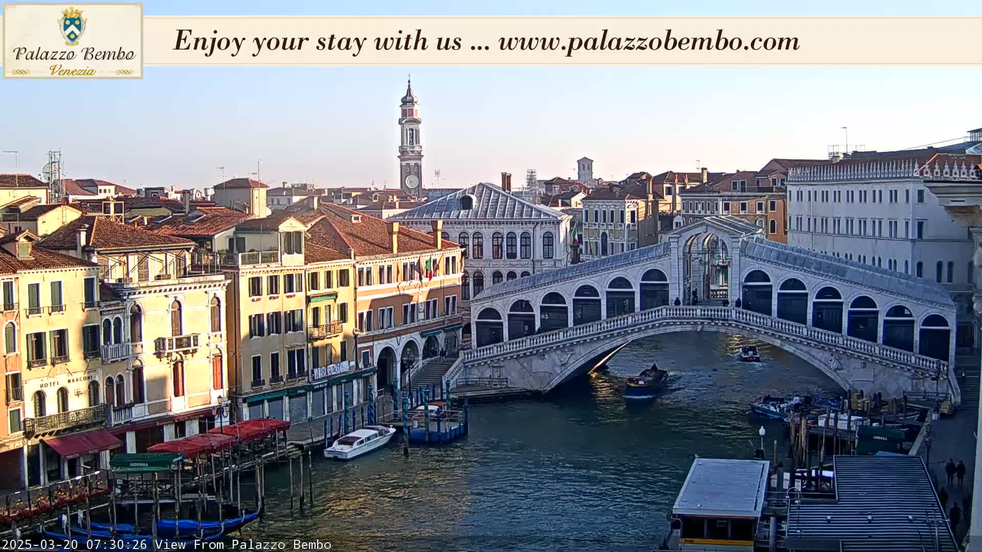 A Venetian canal scene under a clear, light blue sky shows a bridge, numerous buildings, and several boats.