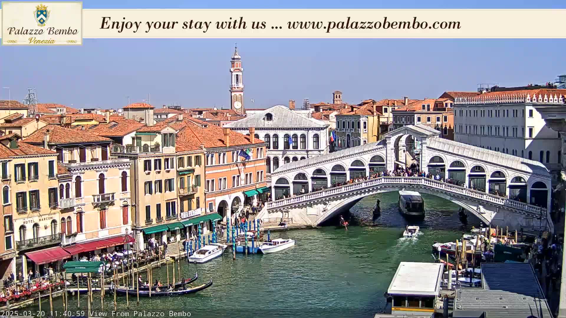 A sunny day in Venice shows the Rialto Bridge bustling with people, gondolas and boats on the canal below, and buildings lining the waterway.
