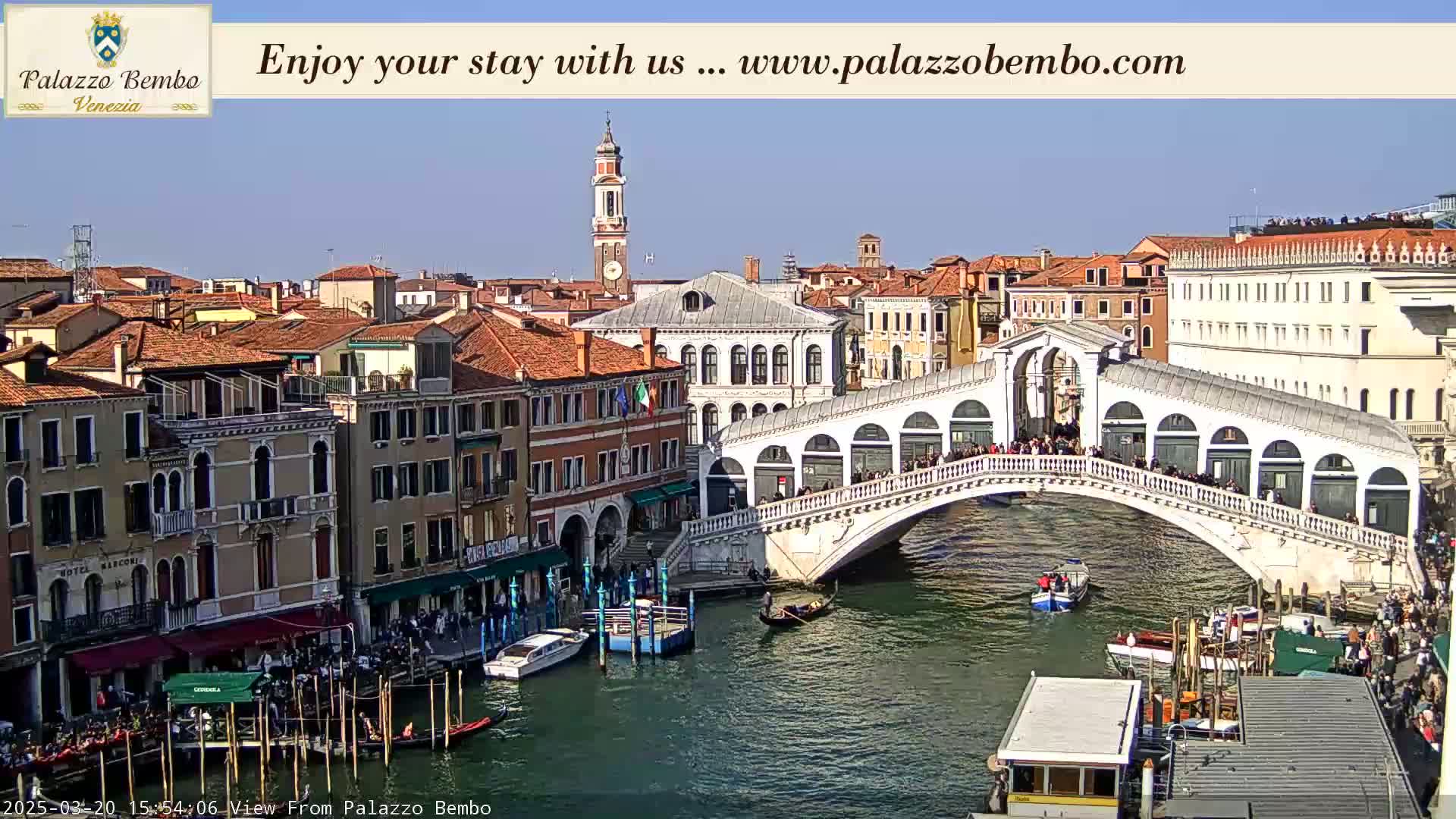 A sunny Venetian canal scene shows a bridge crowded with people, boats on the water, and colorful buildings lining the waterway.