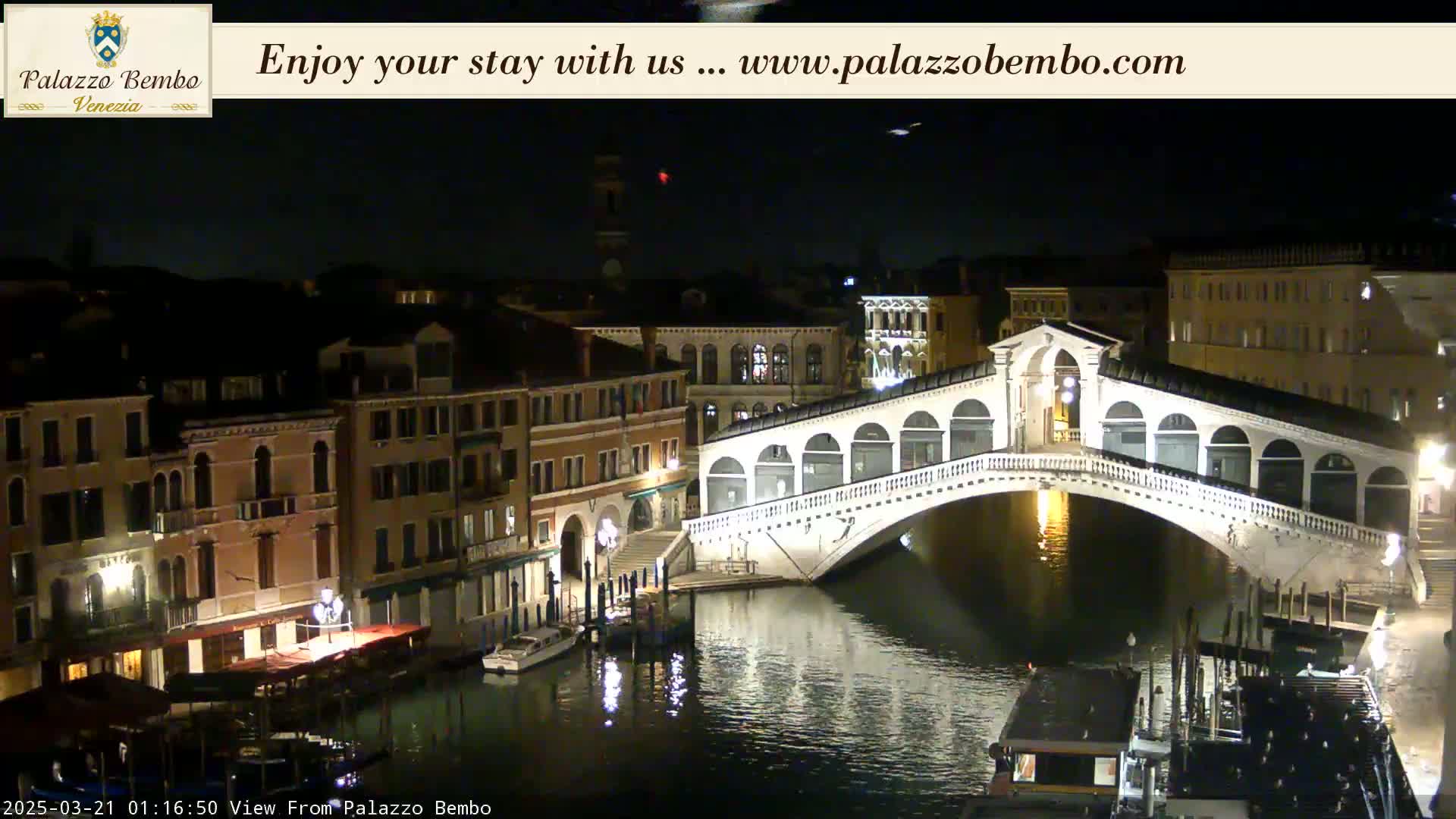 A nighttime view of a bridge over a canal in Venice, with buildings illuminated along the waterway under a clear, dark sky.