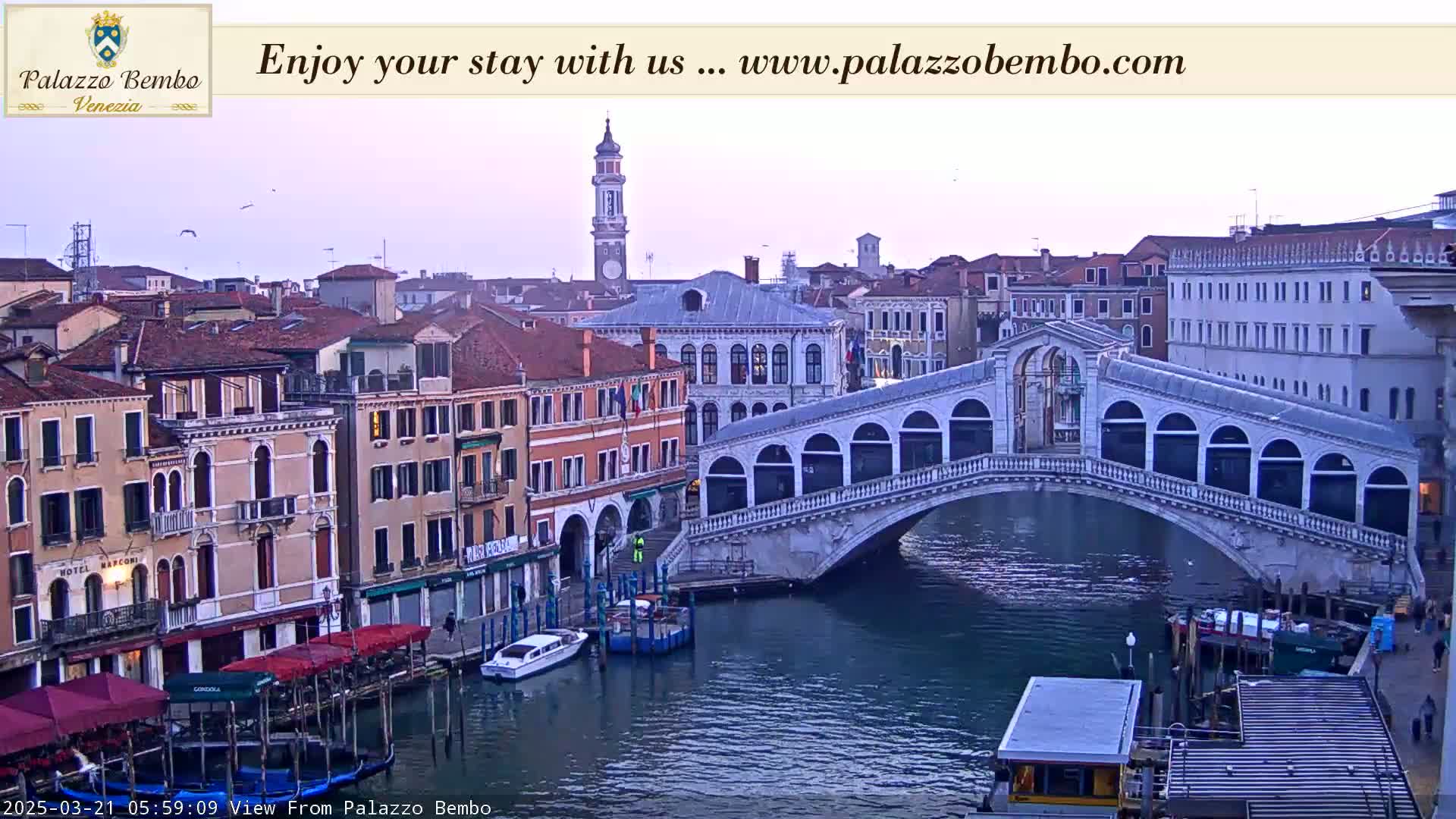 A view of the Rialto Bridge in Venice at dawn, with calm water reflecting the buildings lining the canal.