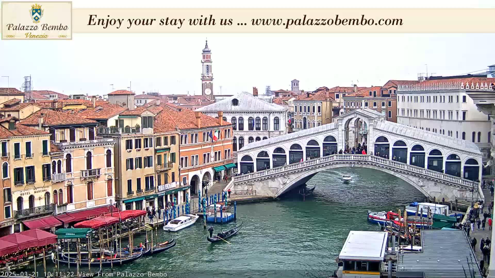 A view of a canal in Venice, Italy, on an overcast day, shows gondolas, boats, and the Rialto Bridge with people walking across it, surrounded by numerous buildings.