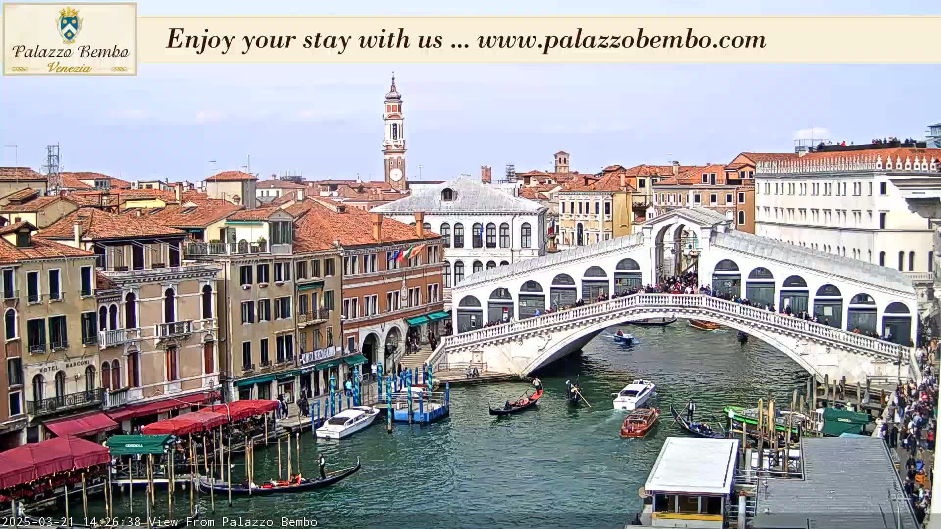 A sunny view of a canal in Venice, showing a bridge crowded with people, gondolas in the water, and numerous buildings lining the waterway.