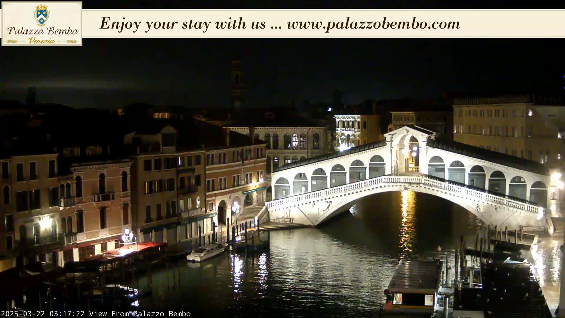 A nighttime view of a canal in Venice, showing the Rialto Bridge illuminated and reflected in the dark water, with buildings lining the waterway.