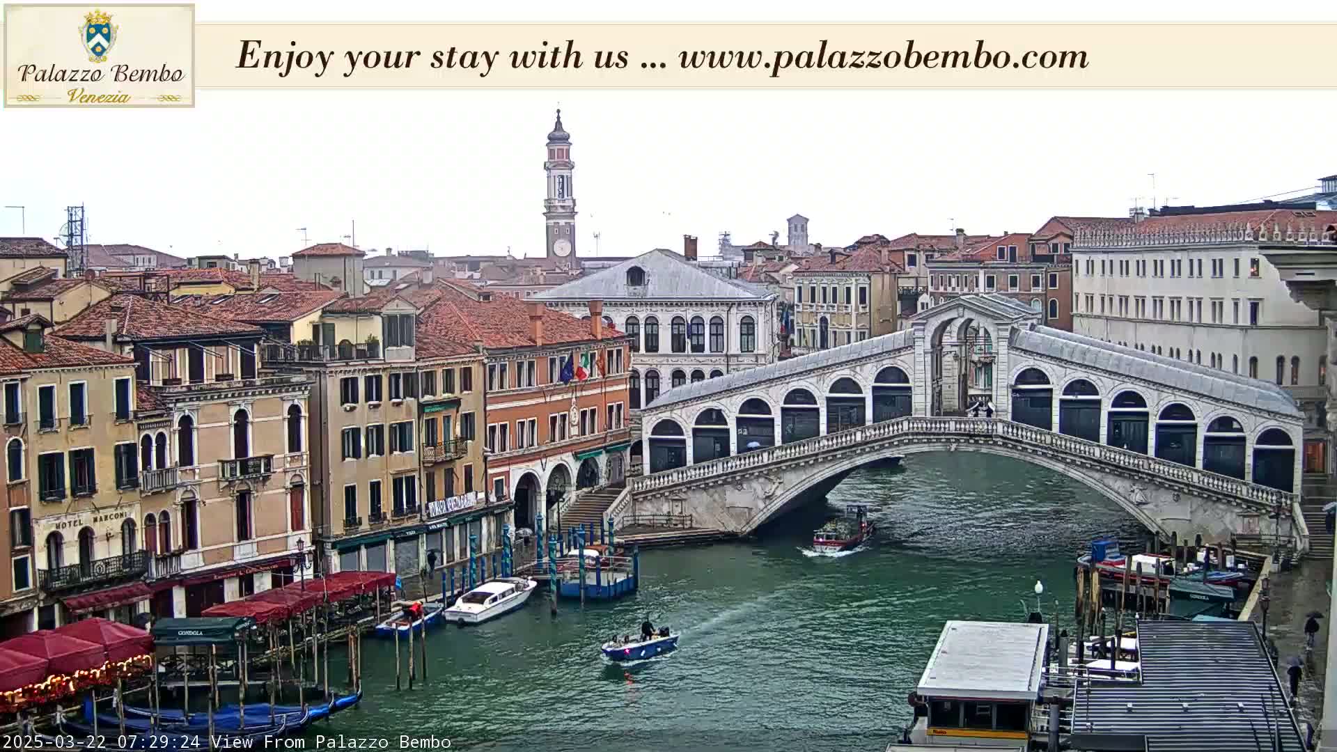 A view of a canal in Venice on an overcast day, showing a bridge with several boats and buildings lining the waterway.