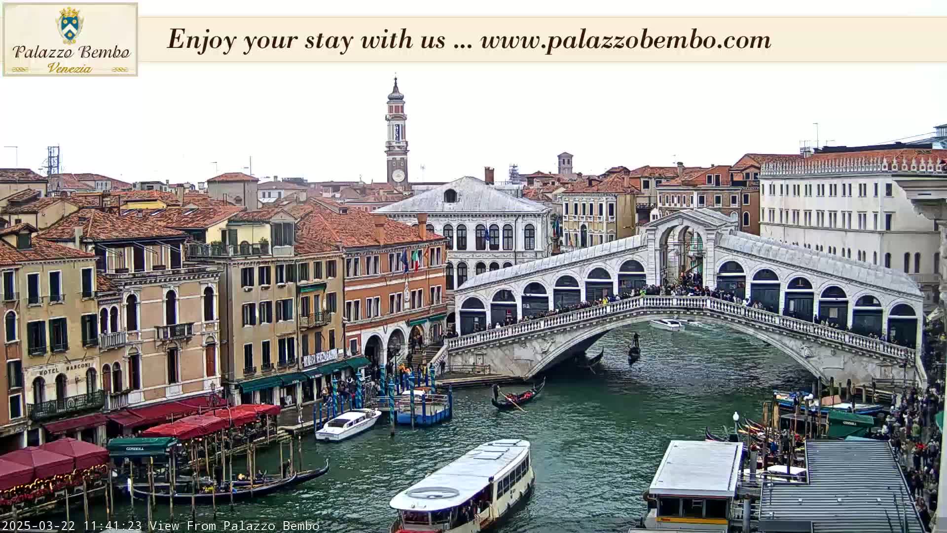 A canal in Venice, under an overcast sky, shows a bridge crowded with people, gondolas, and water taxis.