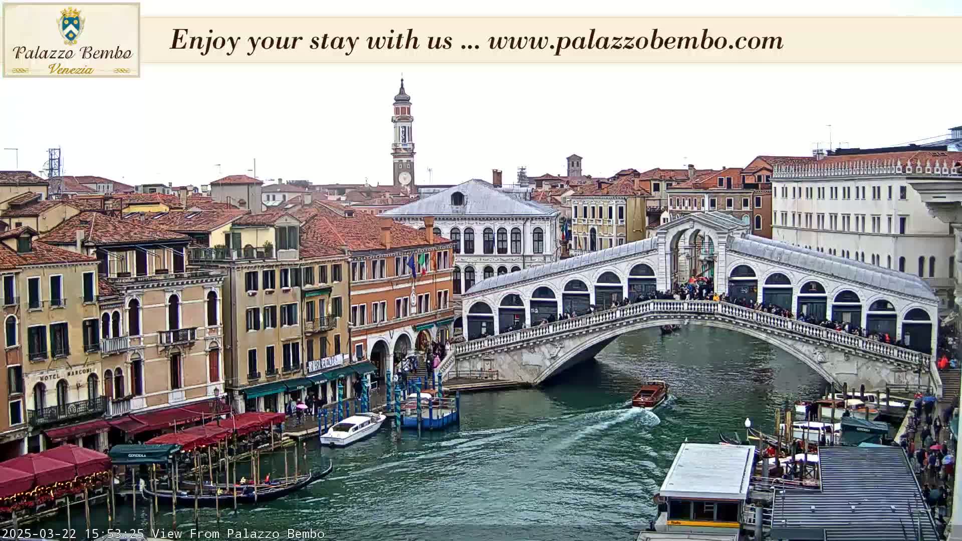 A canal in Venice, under an overcast sky, shows a bridge crowded with people, boats on the water, and buildings lining the waterway.
