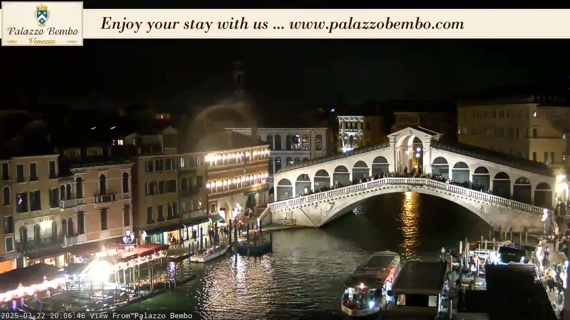 A nighttime view of a bridge in Venice, with many people on it and boats in the canal below, shows some buildings with lights on.