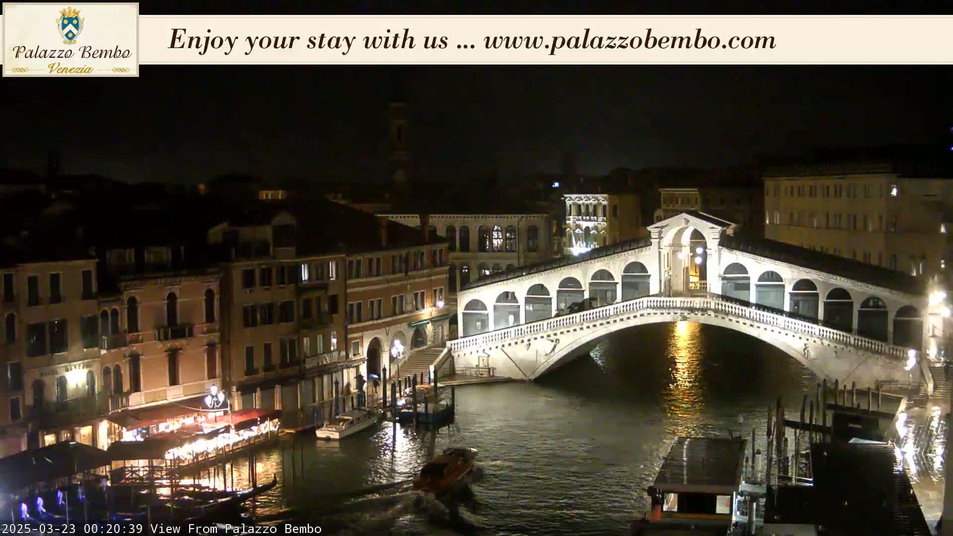 A nighttime view of the Rialto Bridge in Venice, with boats on the canal and illuminated buildings lining the waterway.