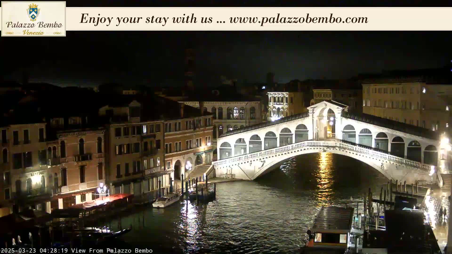 A nighttime view of a canal in Venice, showing a bridge illuminated against a dark sky.