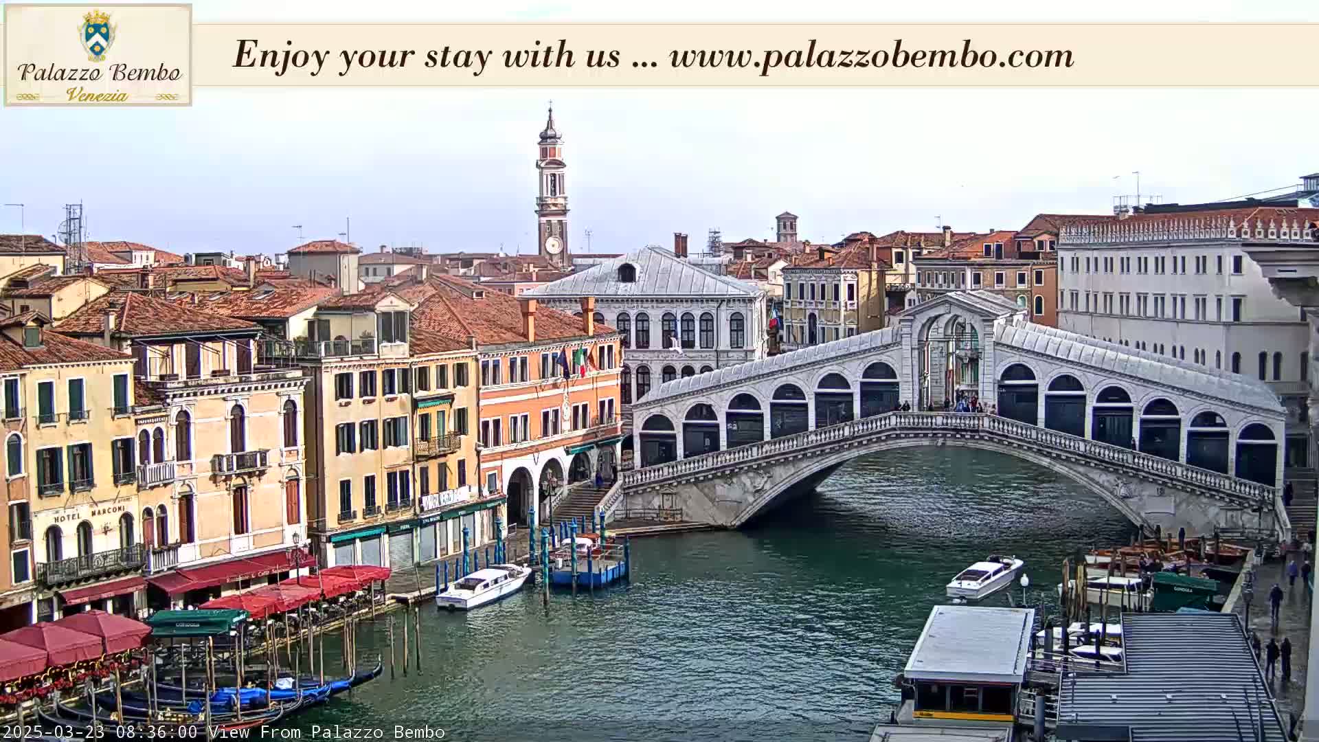 A view of a canal in Venice, Italy, on a partly cloudy day, shows a stone bridge with people walking across it, gondolas and other boats moored along the canal sides, and numerous buildings lining the waterway.
