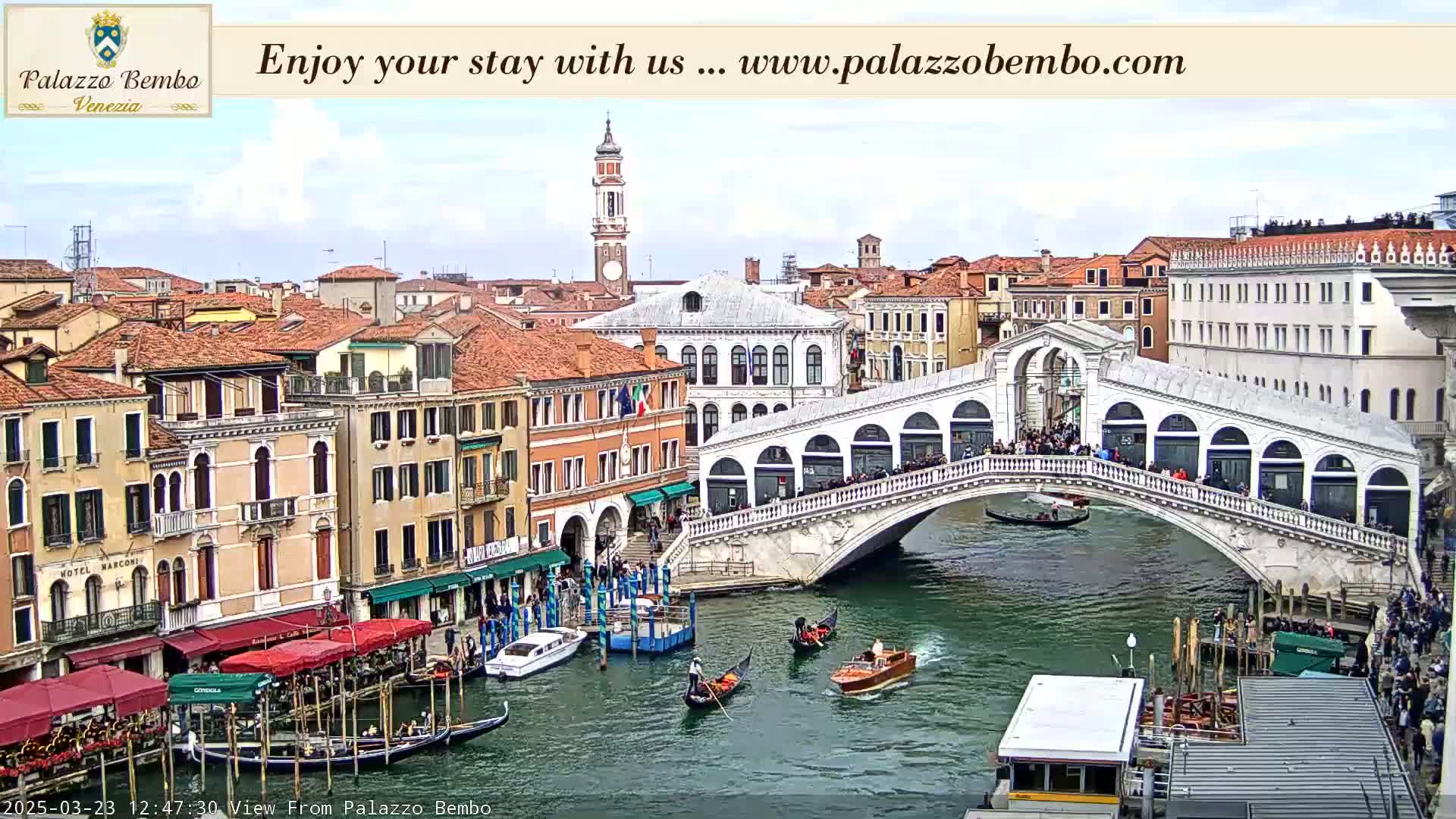 A Venetian canal scene on a partly cloudy day shows a bridge crowded with people, gondolas on the water, and buildings lining the waterway.