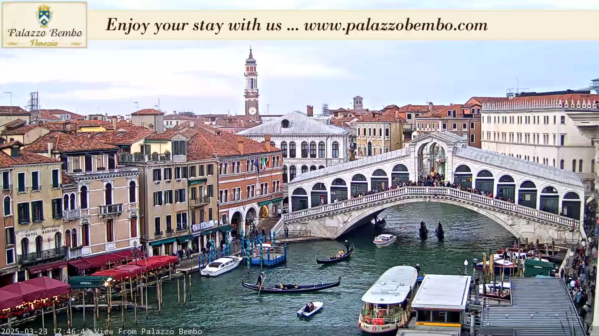 A Venetian canal scene on a partly cloudy day shows a bridge crowded with people, gondolas, and water taxis on the water below, with numerous buildings lining the canal.