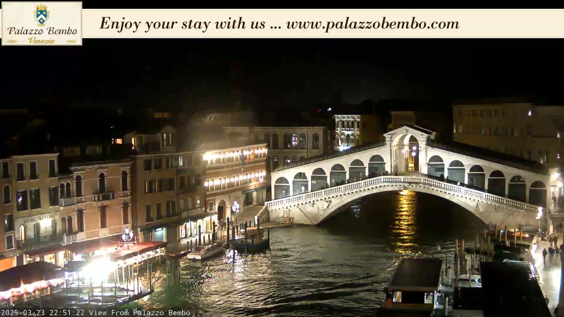 A nighttime view of a canal in Venice, featuring the Rialto Bridge illuminated, with boats and buildings along the waterway.