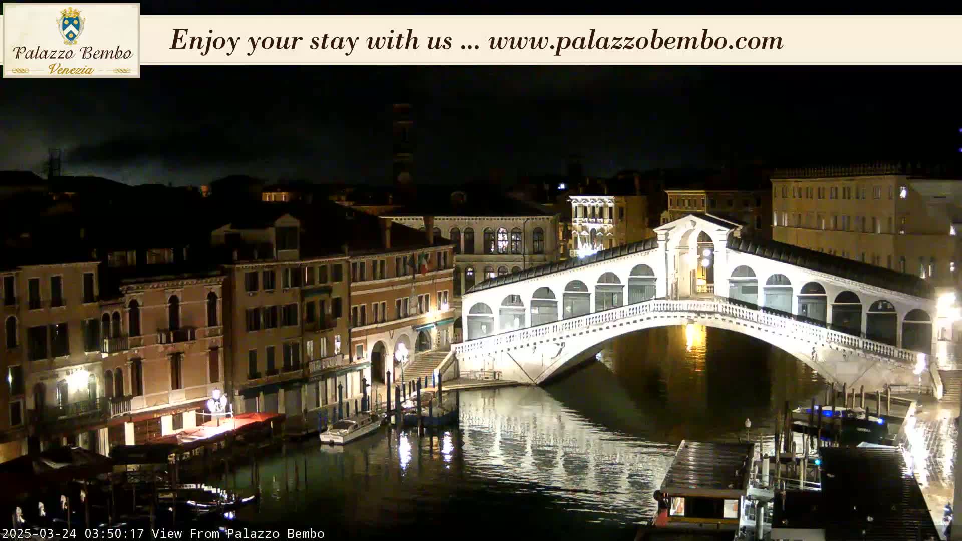 A nighttime view of a canal in Venice, showing a white bridge illuminated against a dark sky.