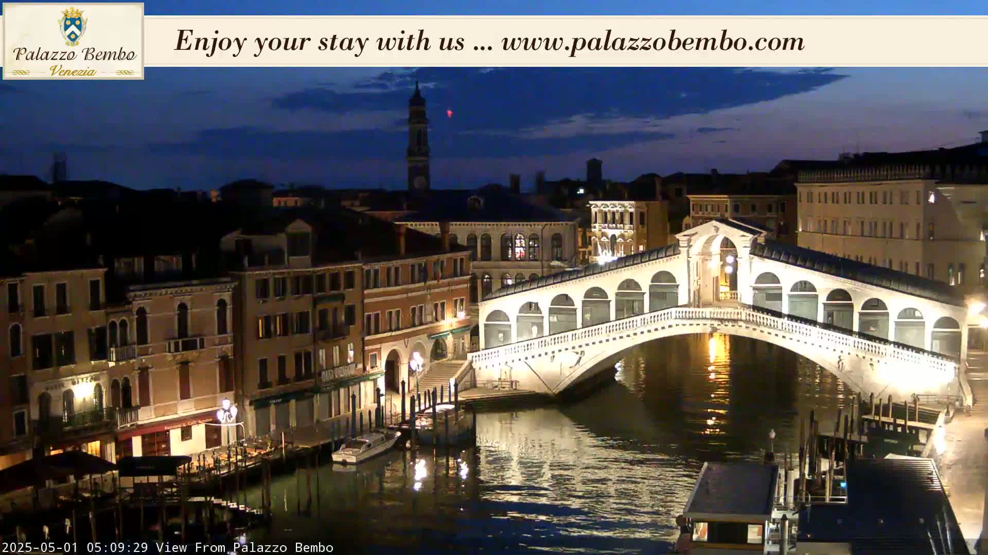 A nighttime view of a canal in Venice, showing a lit bridge and buildings along the water's edge under a dark, mostly clear sky.
