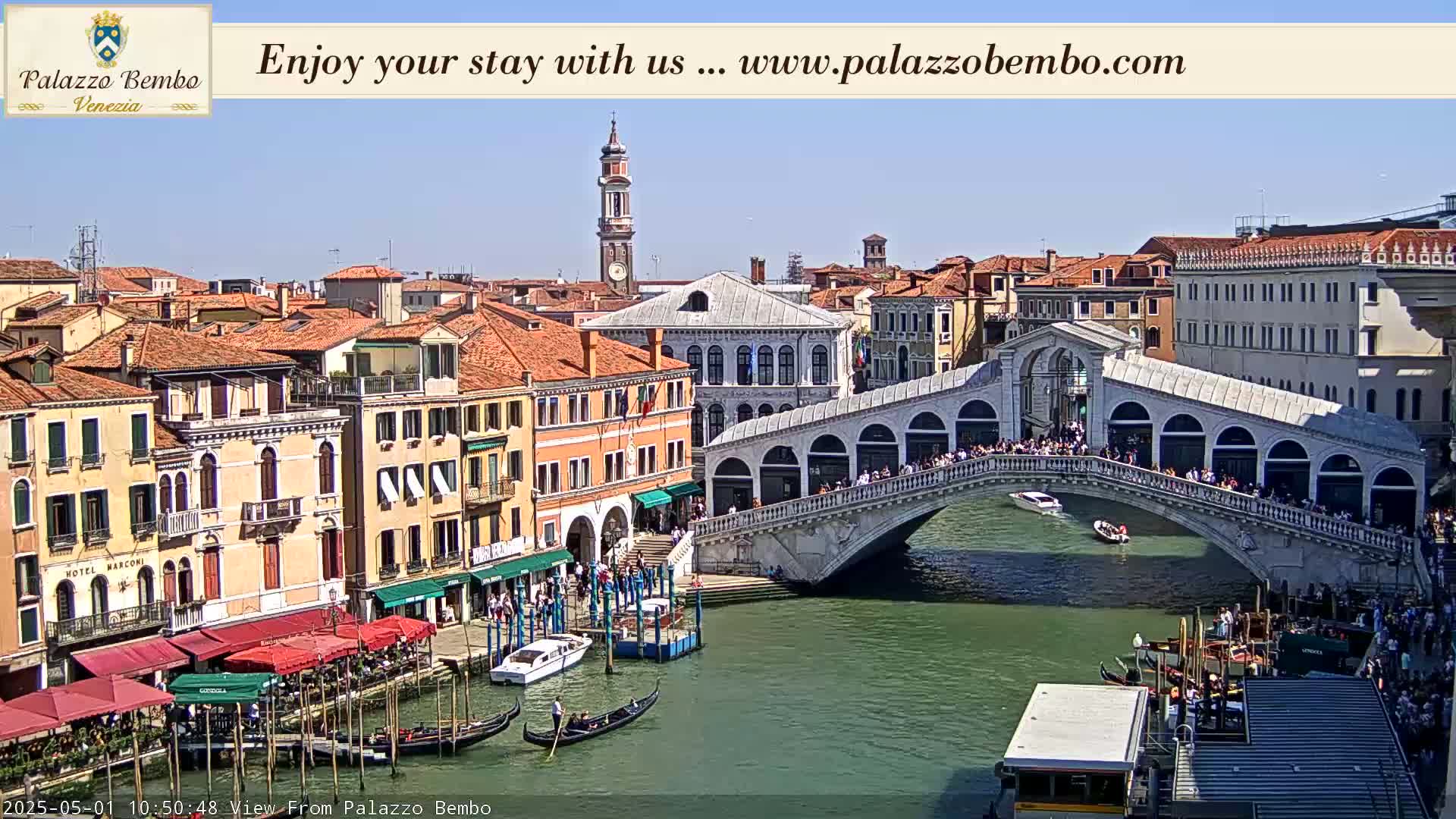 A sunny day in Venice shows a canal with gondolas, boats, and a bridge crowded with people.