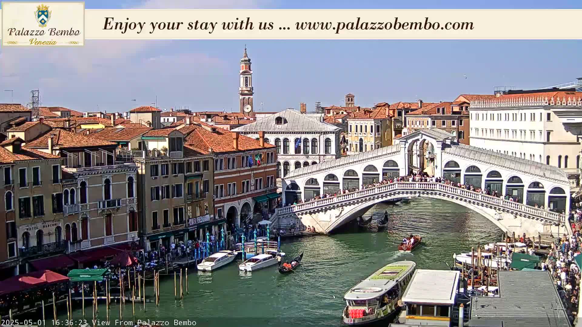 A sunny Venetian cityscape shows the Rialto Bridge crowded with people, gondolas and boats on the canal below, and numerous buildings lining the waterway.