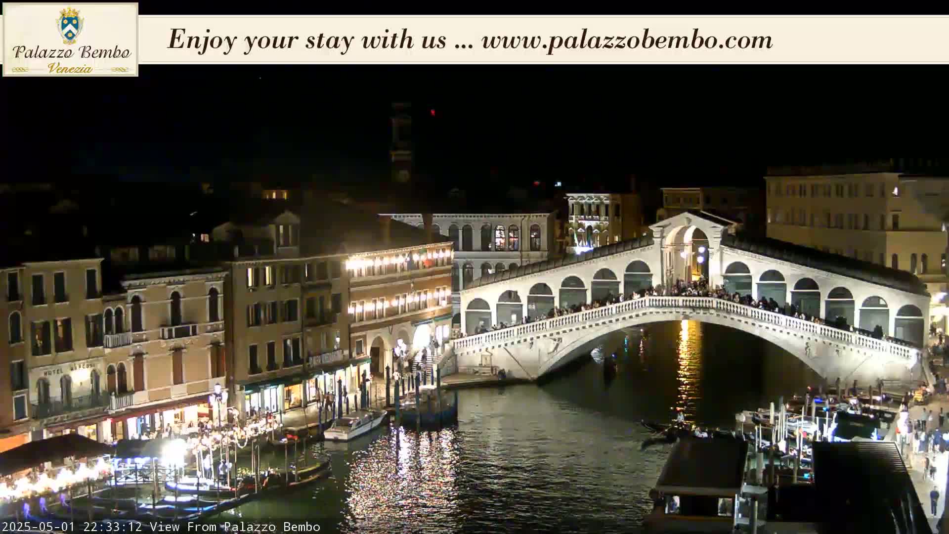 A nighttime view of a crowded bridge over a canal in Venice, with illuminated buildings lining the waterway.