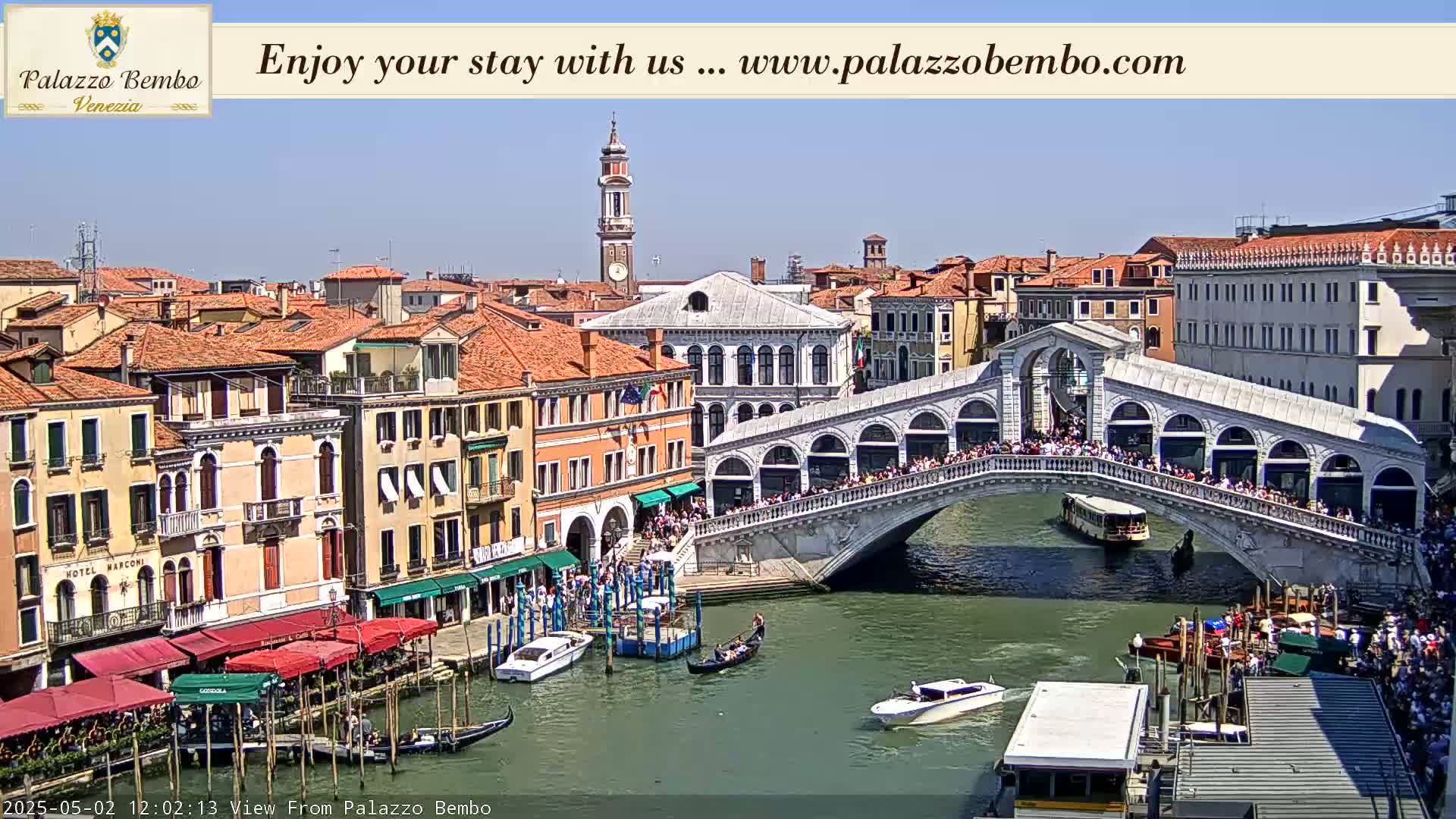 A sunny day in Venice shows a canal with boats, a crowded bridge, and colorful buildings lining the waterway.
