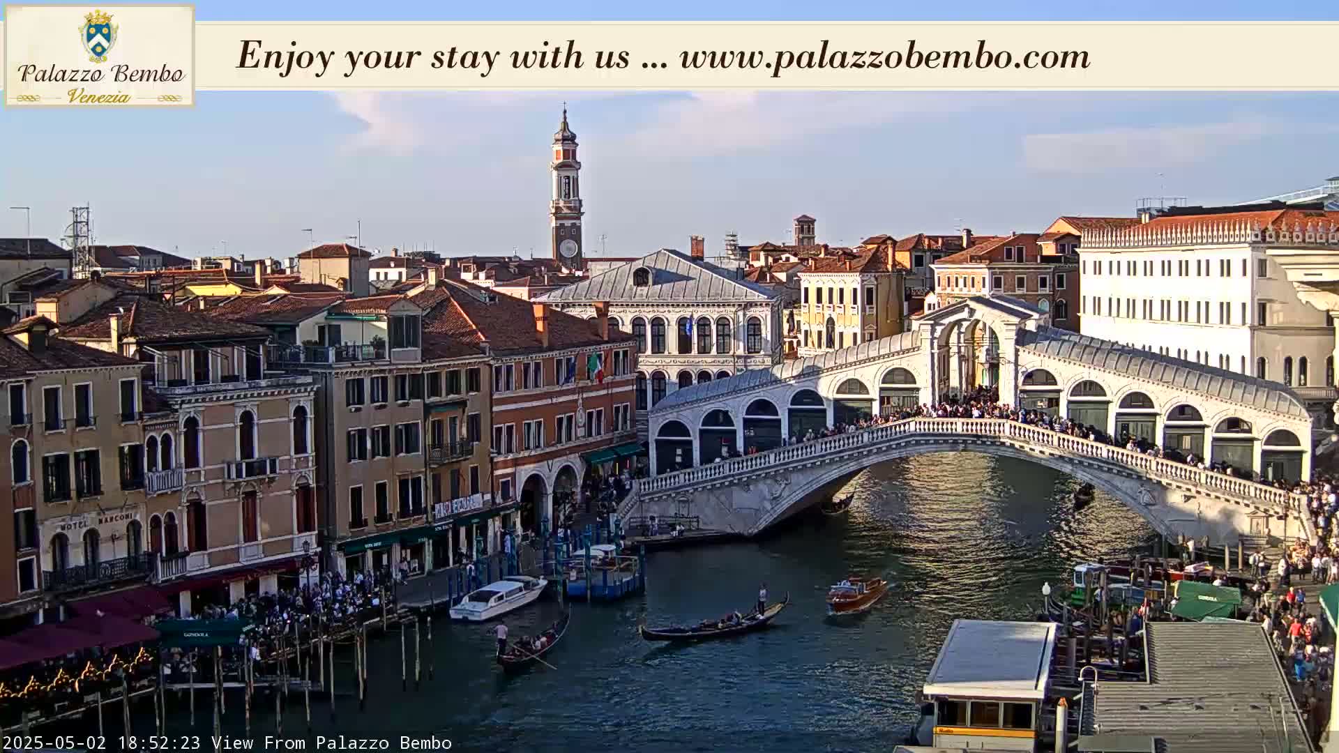 A sunny Venetian canal scene shows a bridge crowded with people, gondolas on the water, and numerous buildings lining the waterway.
