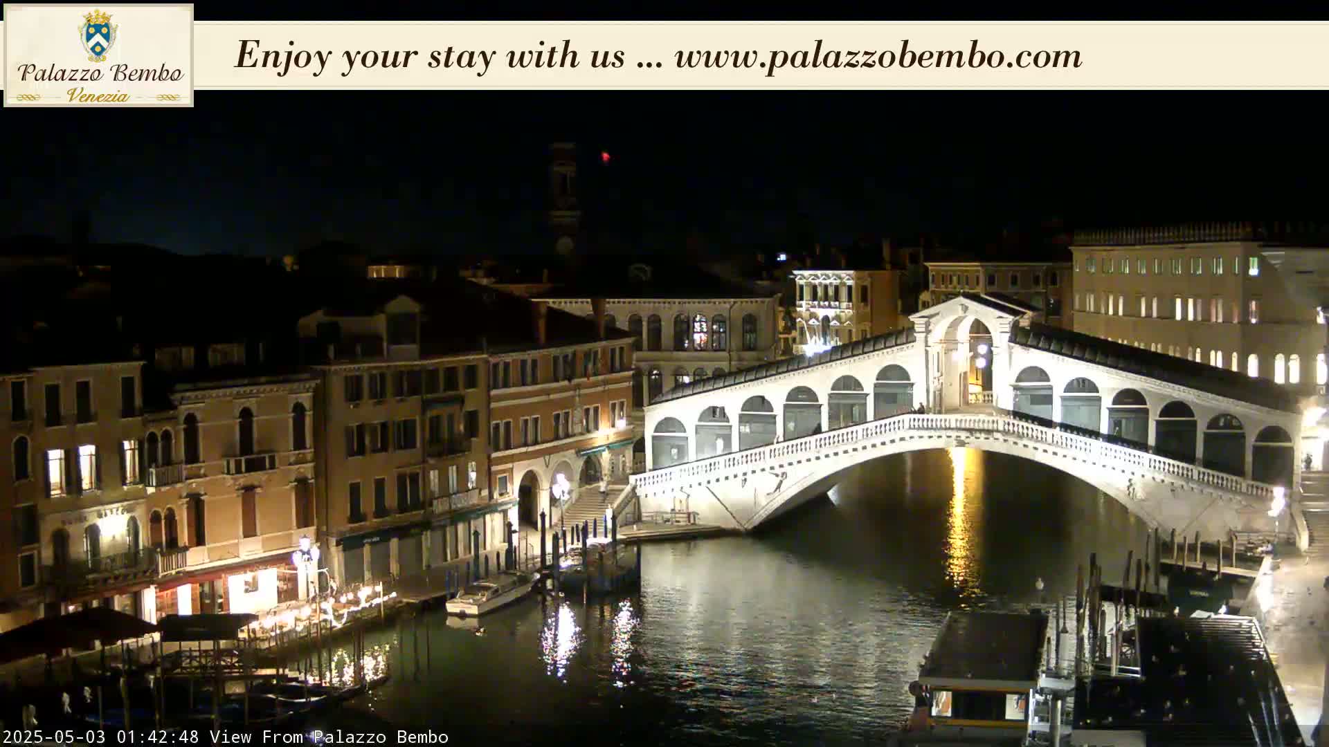 A nighttime view of a canal in Venice, showing a bridge illuminated against a dark sky.