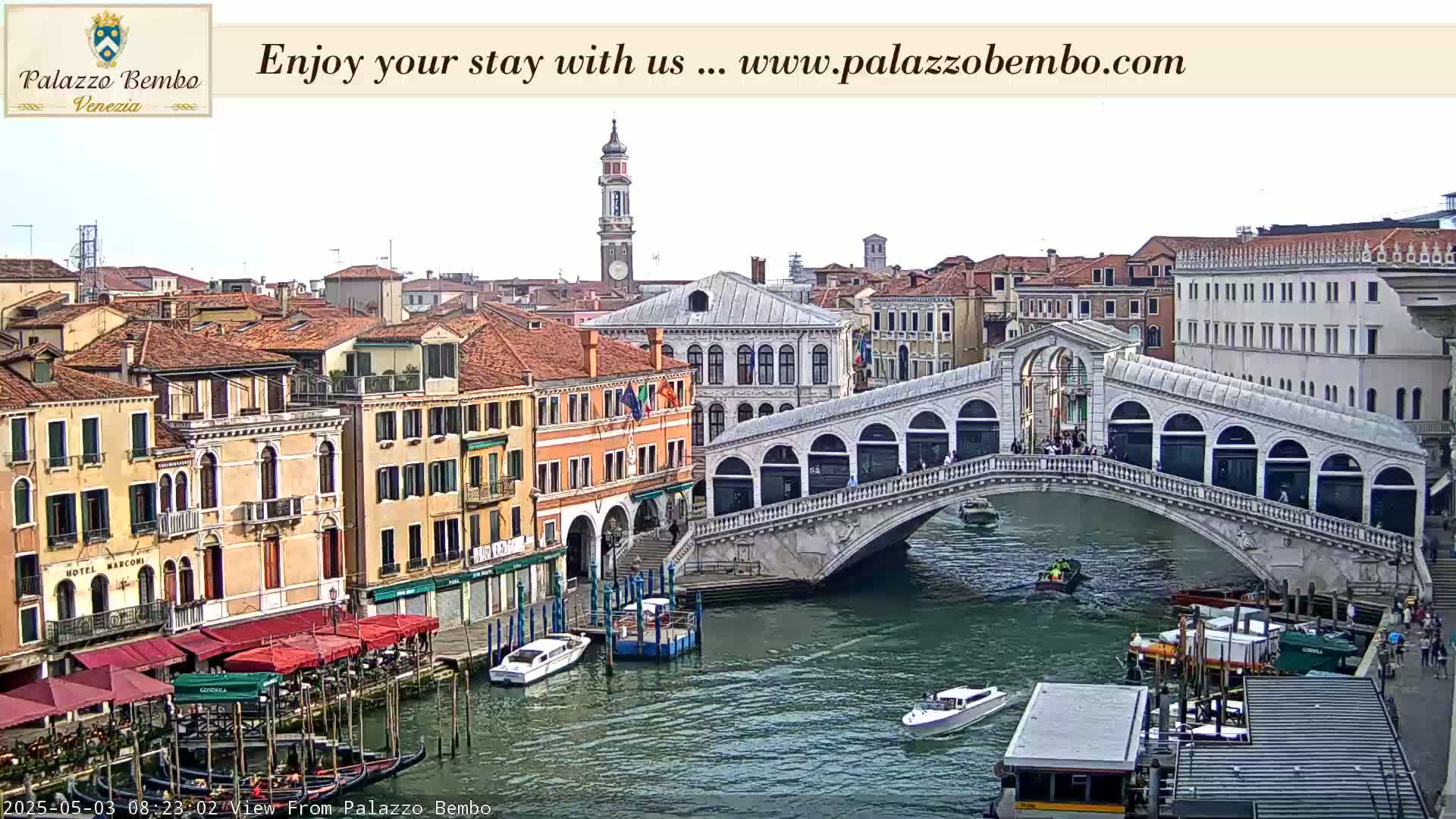 A view of a canal in Venice, under an overcast sky, shows a bridge with people walking across it and boats on the water below, with buildings lining the canal.