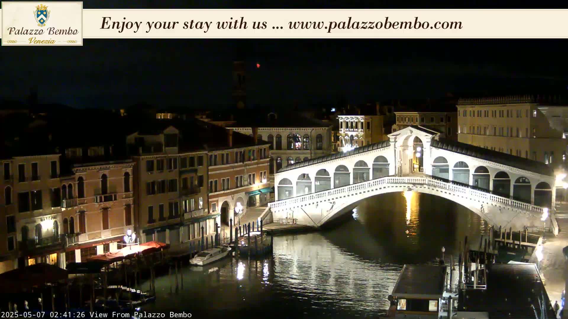 A nighttime view of a canal in Venice, showing a lit bridge and buildings along the waterway under a clear, dark sky.