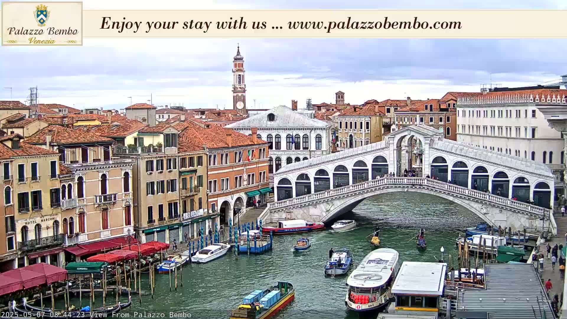 A view of a Venetian canal with boats and the Rialto Bridge, under a partly cloudy sky.