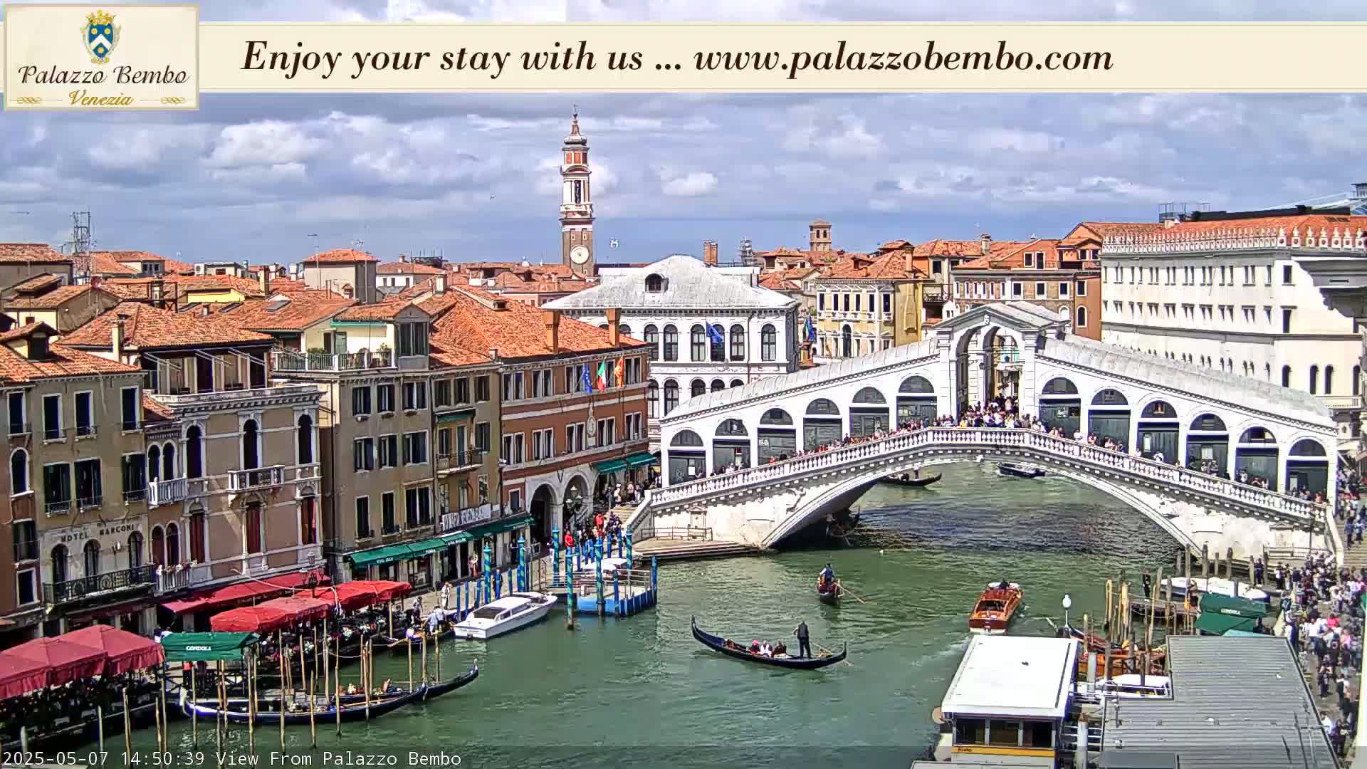A partly cloudy day in Venice shows a canal with gondolas, boats, and a bridge crowded with people, all surrounded by buildings.