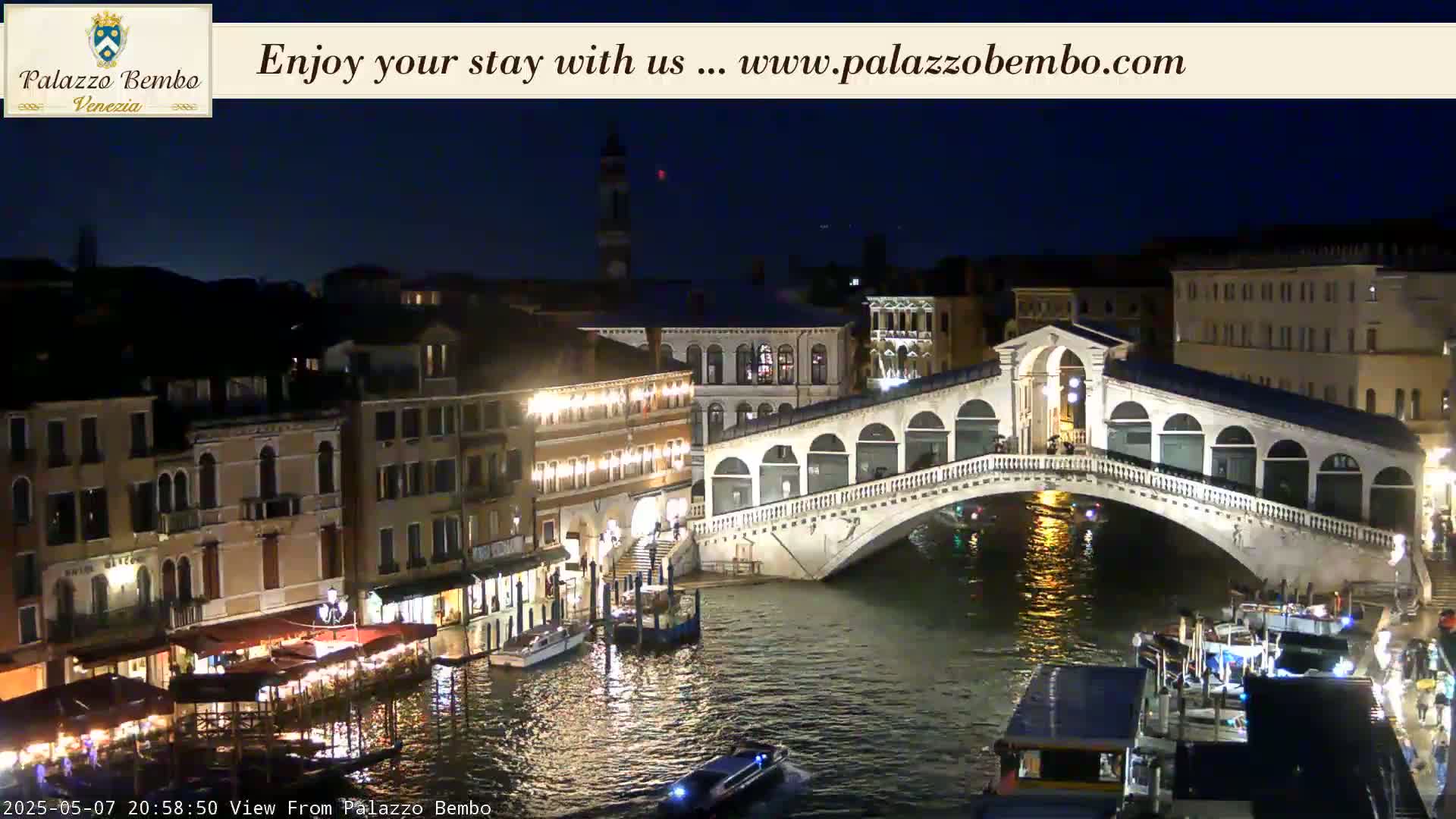 A nighttime view of a canal in Venice, showing a bridge lit up with lights, boats on the water, and buildings along the canal.