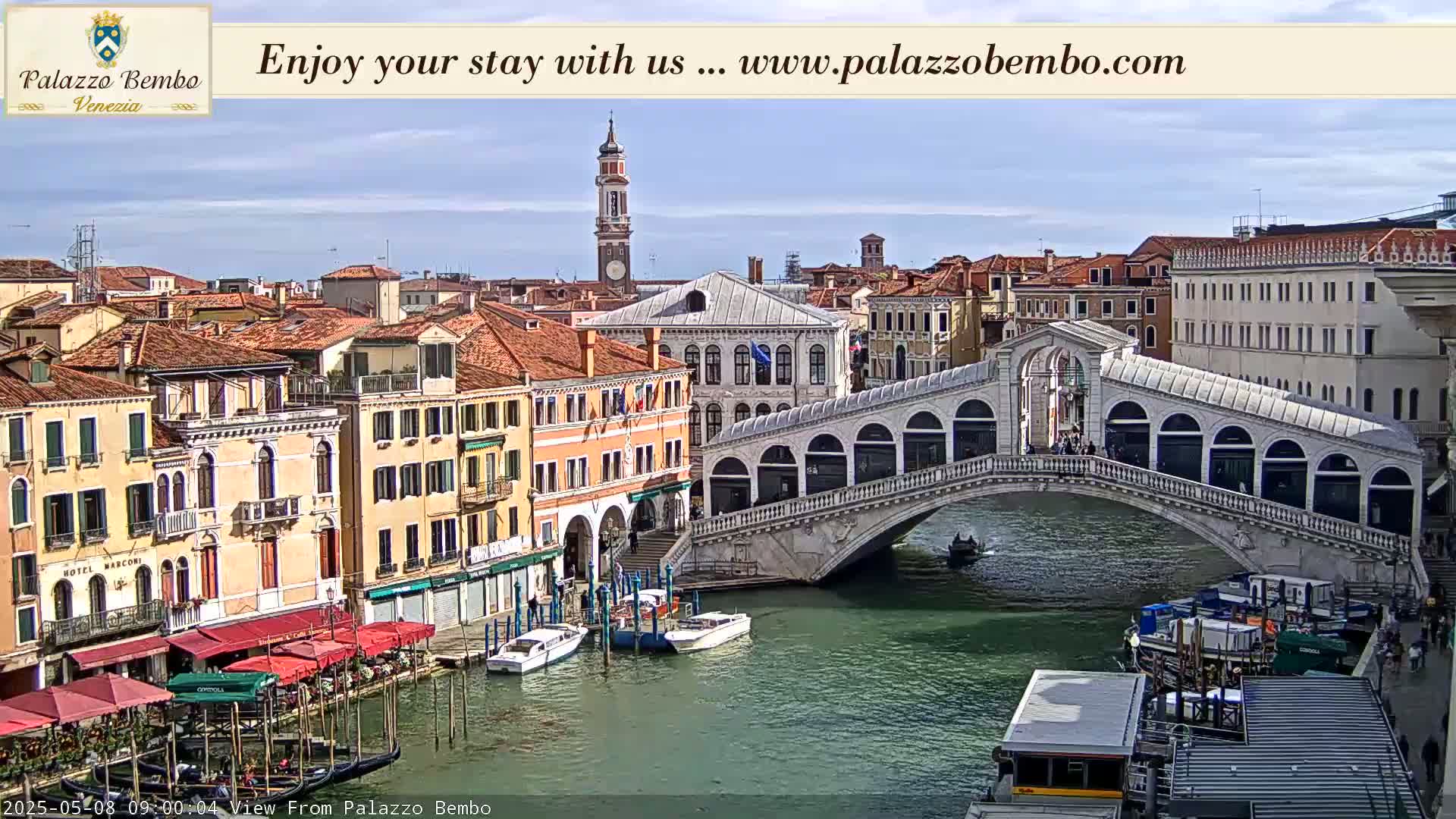A sunny view of a canal in Venice, showing the Rialto Bridge, gondolas, boats, and several buildings.