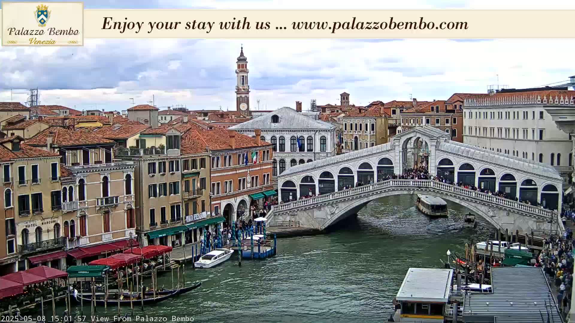A view of the Rialto Bridge in Venice, Italy, on a partly cloudy day, shows many people walking across the bridge and gondolas on the canal below.