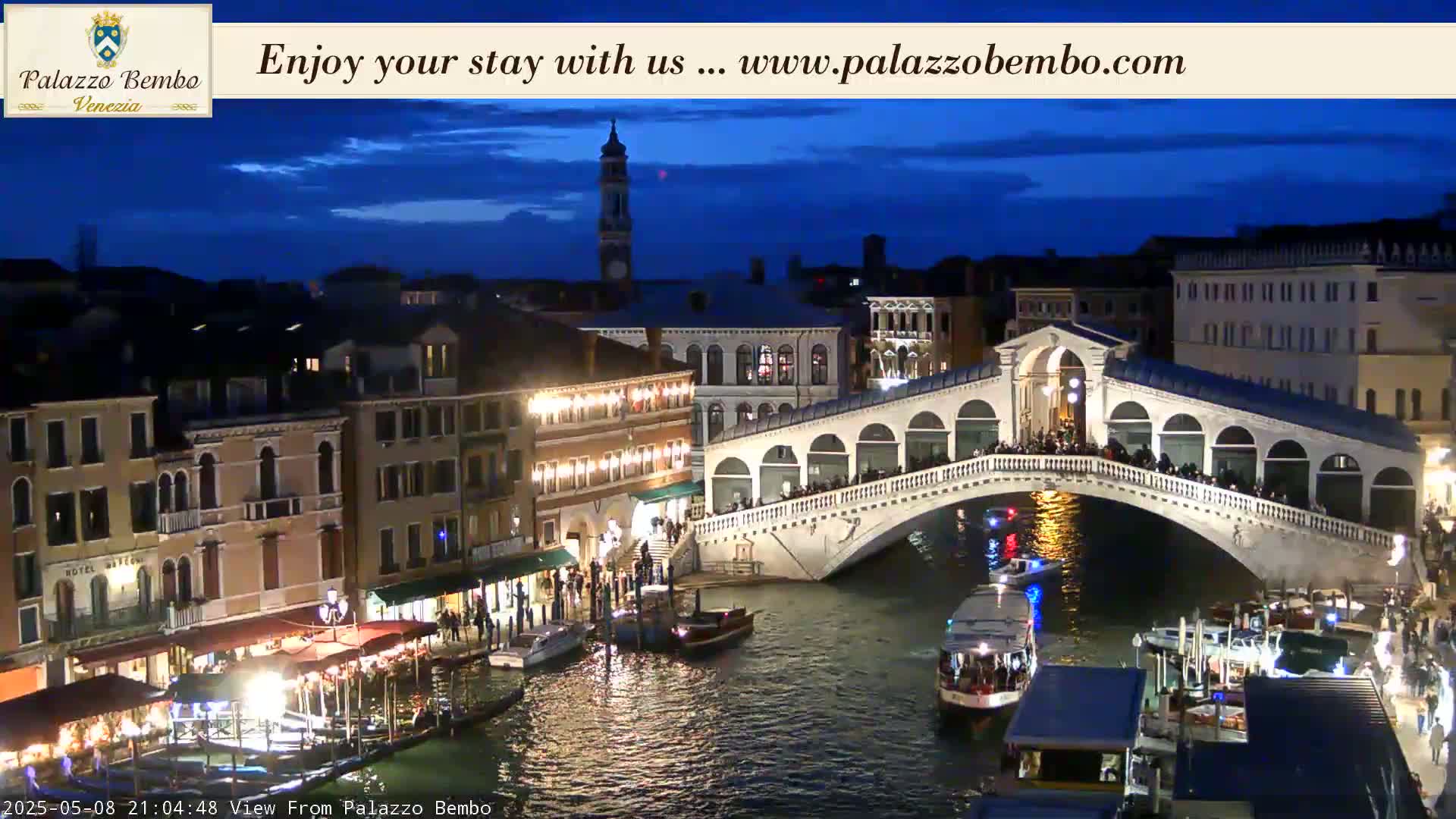 A nighttime view of a canal in Venice, showing a bridge crowded with people, boats on the water, and buildings illuminated along the waterway under a twilight sky.