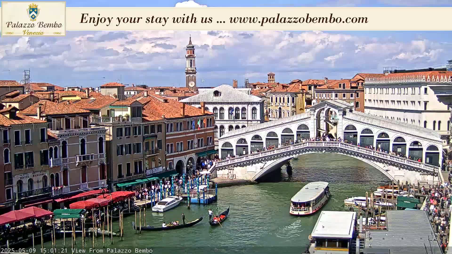 A partly cloudy sky overlooks a Venetian canal, with gondolas and water taxis traversing beneath the Rialto Bridge, lined with people.