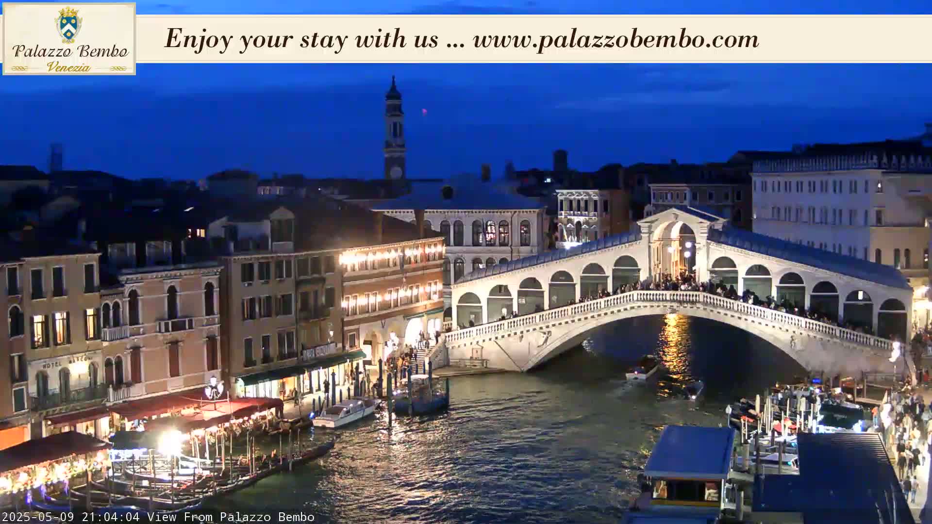 A nighttime view of a bridge over a canal in Venice, with many buildings and boats visible under a dark blue sky.