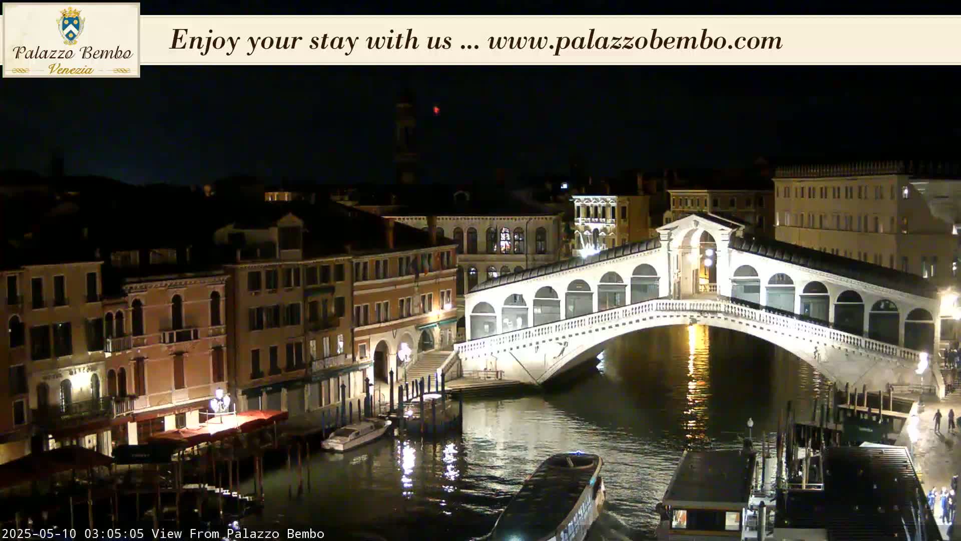 A brightly lit bridge arches over a dark canal at night, with buildings lining the waterway.